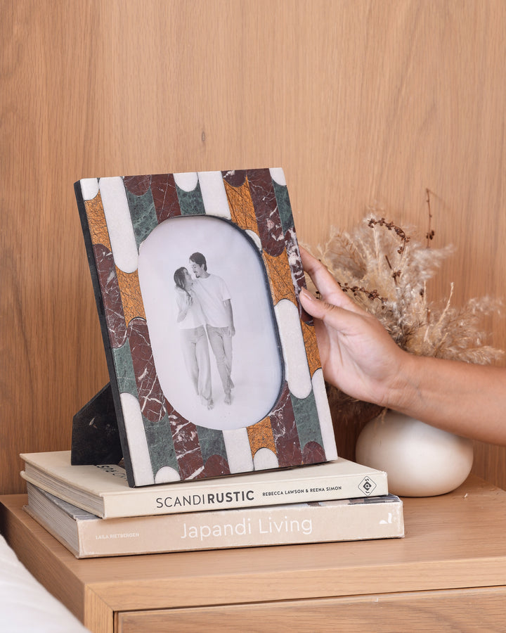 A hand holds a rectangular photo frame with a marbled pattern in brown, green, and cream, featuring an oval cutout with a couple's silhouette. The frame is displayed on a stack of books on a wooden table next to a vase with dried grass.
