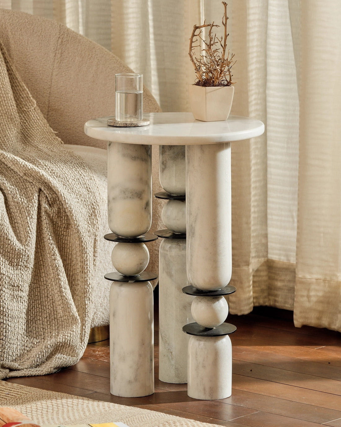 A small round side table with a white marble-look top and sculptural cylindrical legs with dark metal accents, styled in a cozy living room next to a sofa with a textured throw blanket.